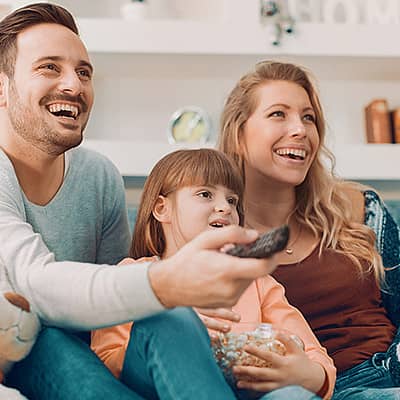 Familie sitzt auf dem Couch im Wohnzimmer und schaut Fern. Der Mann hält die Fernbedienung.